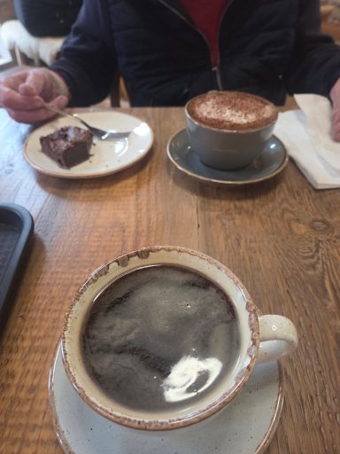 A cup of black coffee, a cappuccino, and a slice of cake on a wooden table.