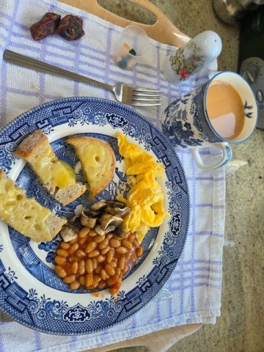 A blue plate with toast, scrambled eggs, beans, and a cup of tea on a cloth-covered table.